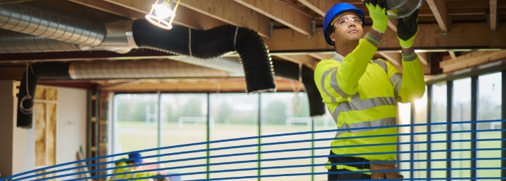 Construction worker in safety gear installing HVAC ductwork in an unfinished building ceiling.